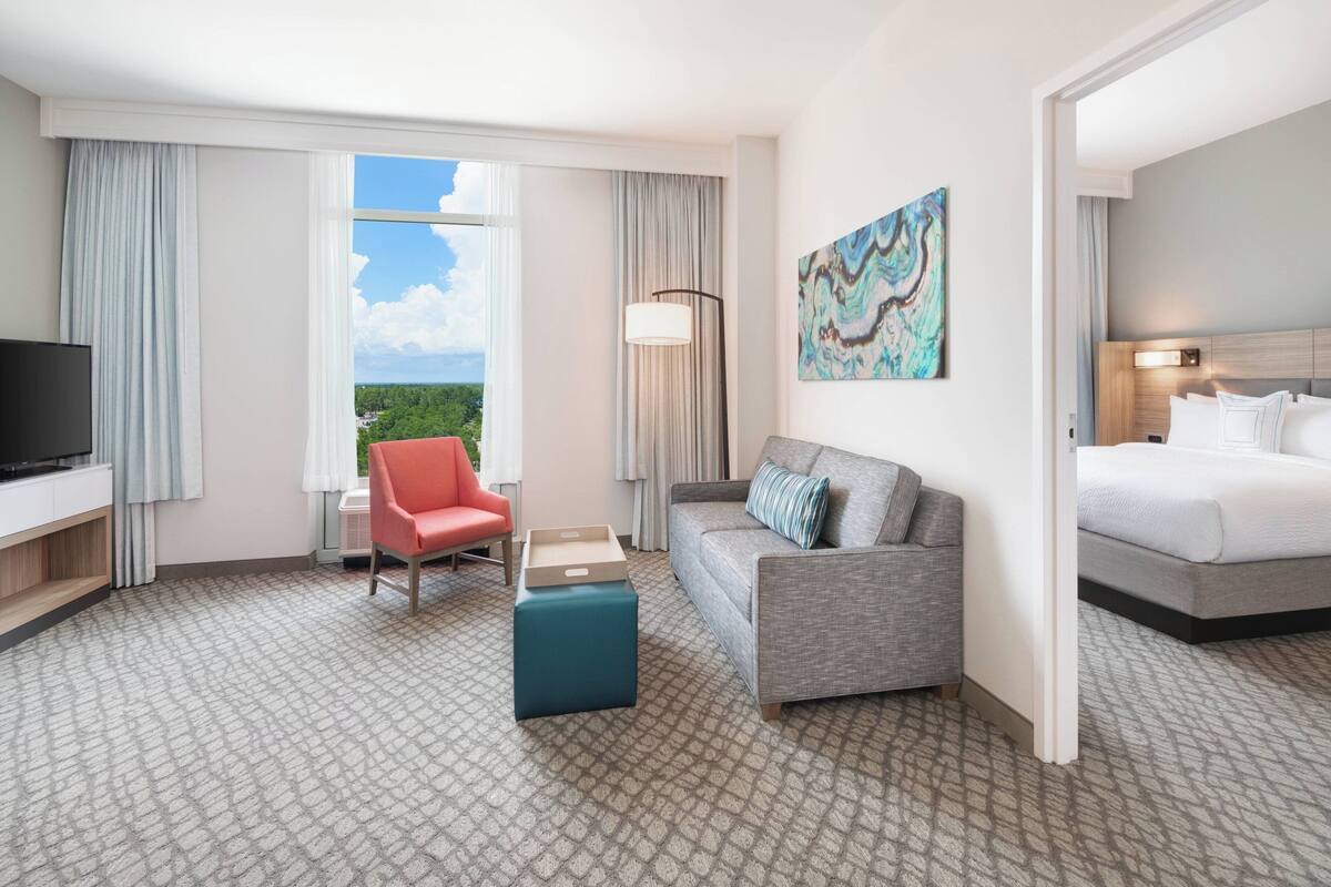 A hotel room with a large window, a sofa, a bed, a TV, and a painting on the wall.