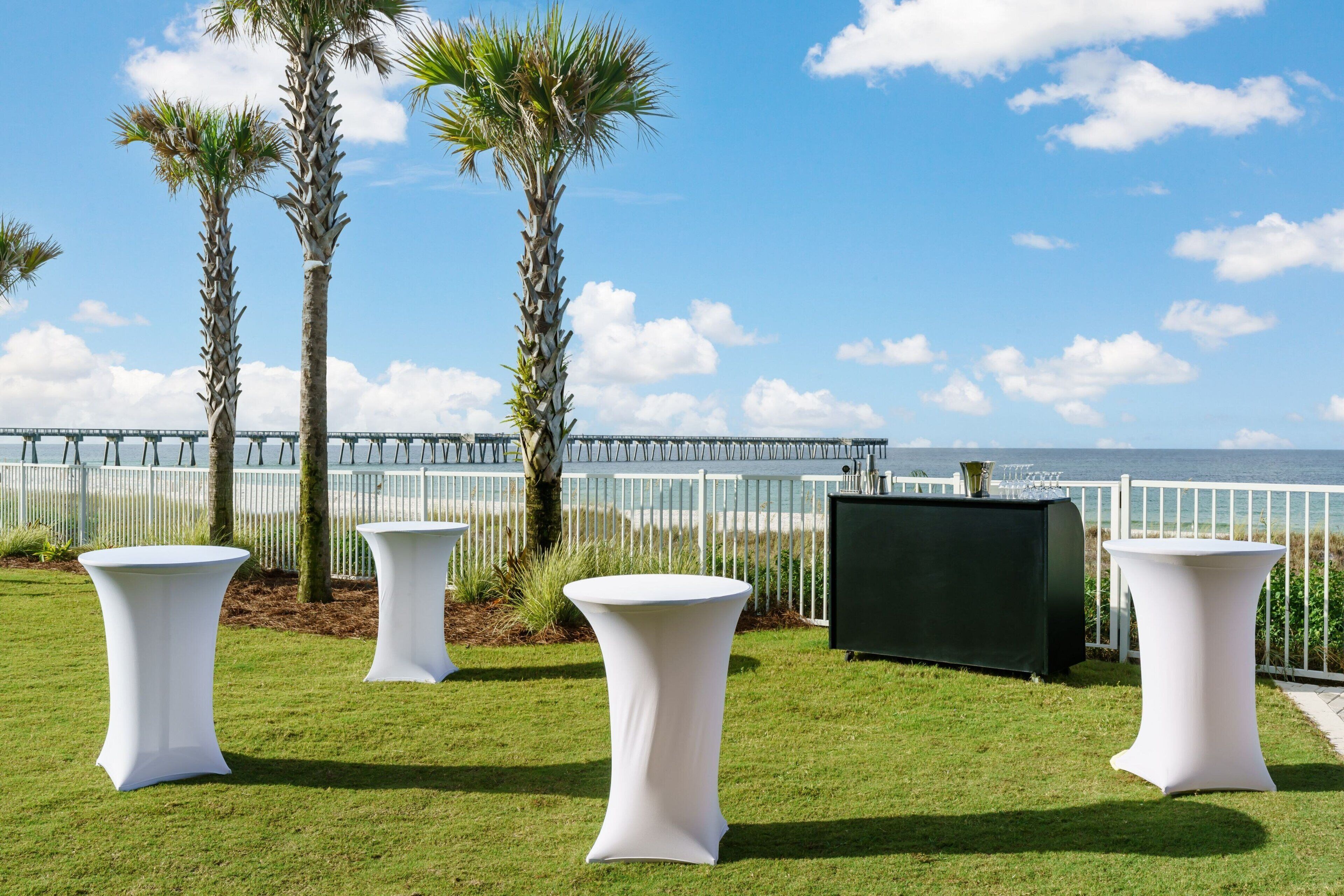 Un espace extérieur verdoyant, avec des tables blanches et un barbecue noir, offrant une vue sur une jetée et la mer.