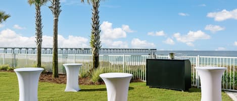 Un espacio al aire libre con césped, mesas blancas y una parrilla negra, con vistas a un muelle y al mar.