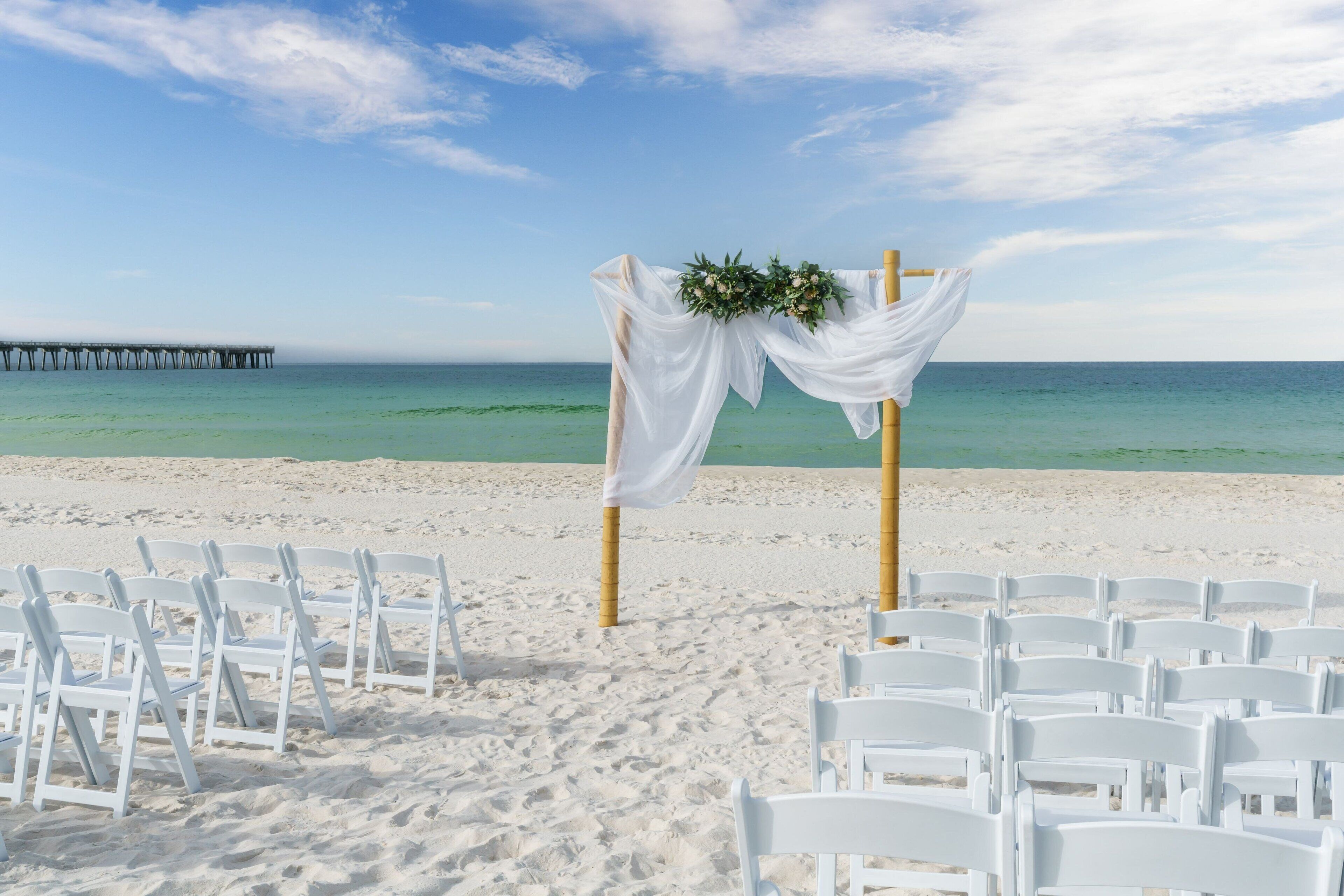 Une cérémonie célébrée sur la plage, avec des chaises blanches et une arche décorée de fleurs.