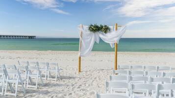 Une cérémonie célébrée sur la plage, avec des chaises blanches et une arche décorée de fleurs.