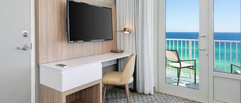 A hotel room with a flat-screen TV mounted on a wooden wall, a desk with a chair, a balcony with a view of the ocean, and a patterned floor.