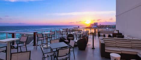 Una terraza en la azotea con mesas y sillas, con vistas al océano y a un atardecer.