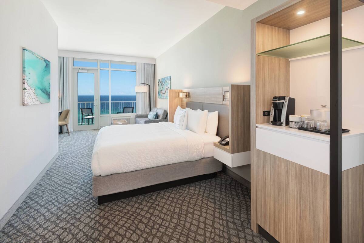 A hotel room with a large bed, a desk with a coffee maker, and a view of the ocean through the window.