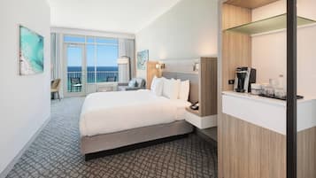 Habitación de hotel con una cama grande, un escritorio con cafetera y vista al océano por la ventana.