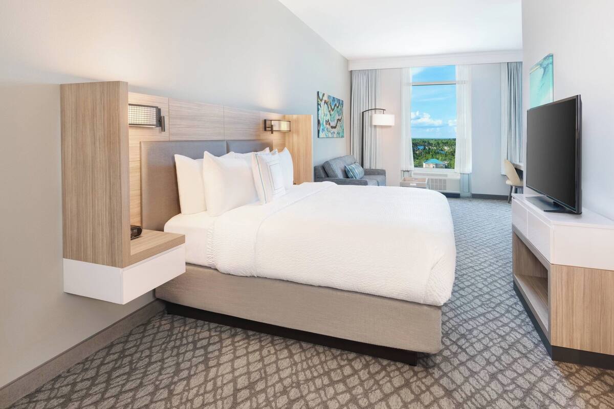 A modern hotel room with a large bed, a TV, a desk, and a view of the outdoors.