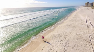 Una playa con una persona caminando por la orilla, cielo despejado y edificios al fondo.