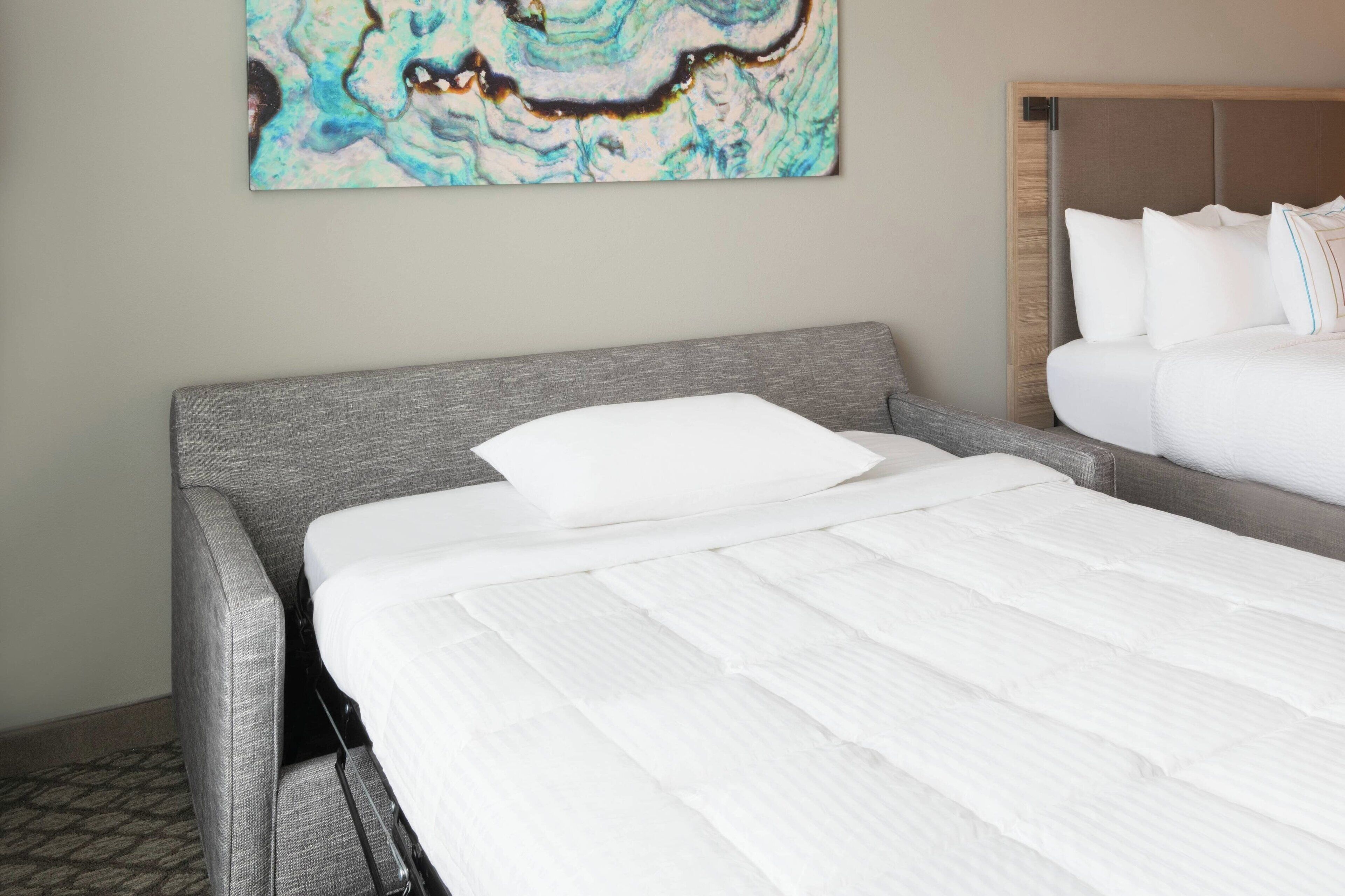 Une chambre d’hôtel avec un lit recouvert d’un tissu gris, un matelas blanc et un grand tableau abstrait accroché au mur.