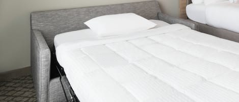 Hotellihuone, jossa on harmaalla kankaalla verhoiltu sänky, valkoinen patja ja suuri abstrakti maalaus seinällä.