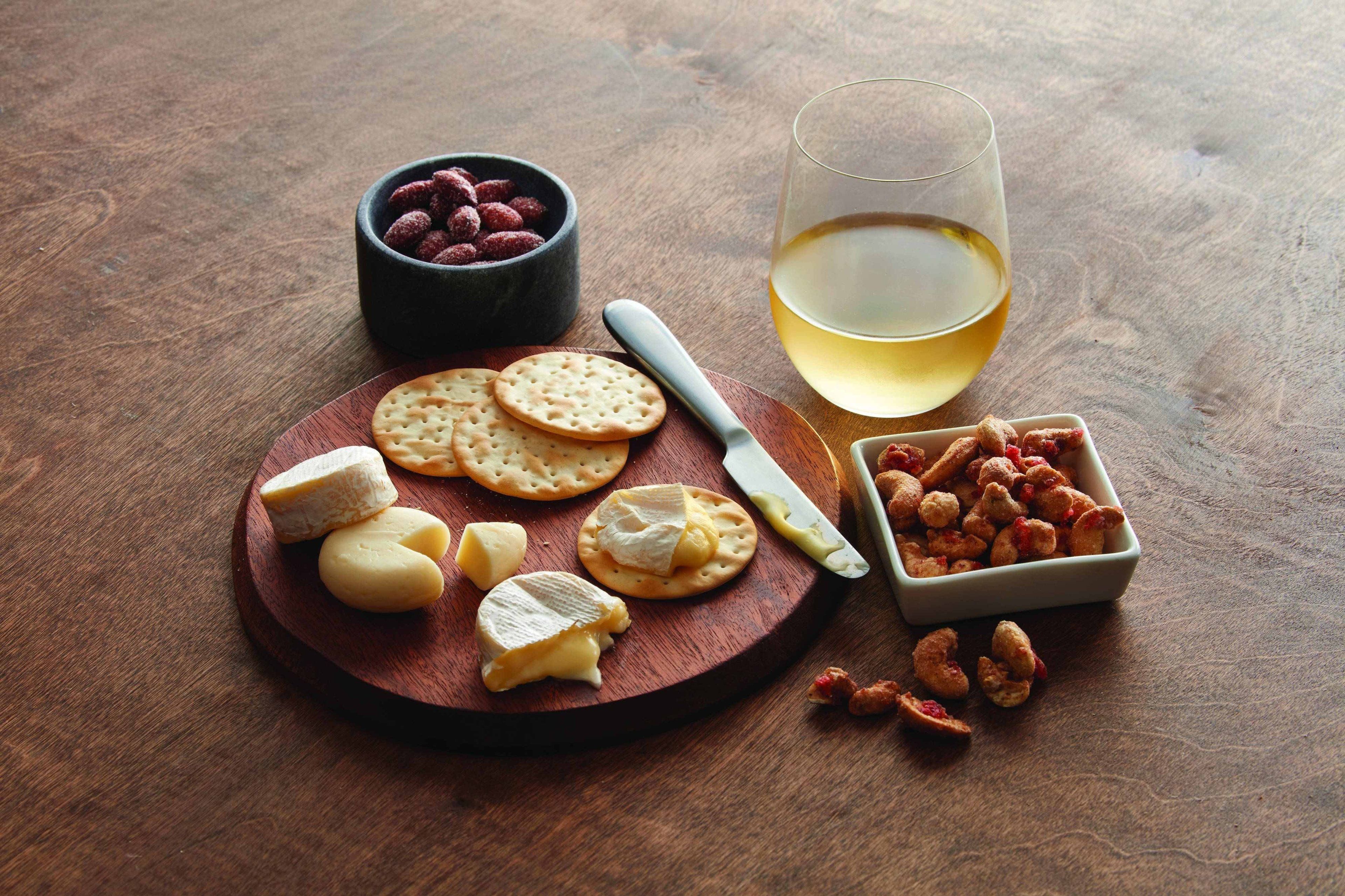 Ett träbord med ett fat med olika sorters ost, kex, en skål med nötter och ett glas vitt vin.