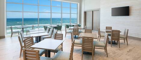 A spacious dining area with large windows overlooking the ocean, wooden furniture, and a flat-screen TV mounted on the wall.