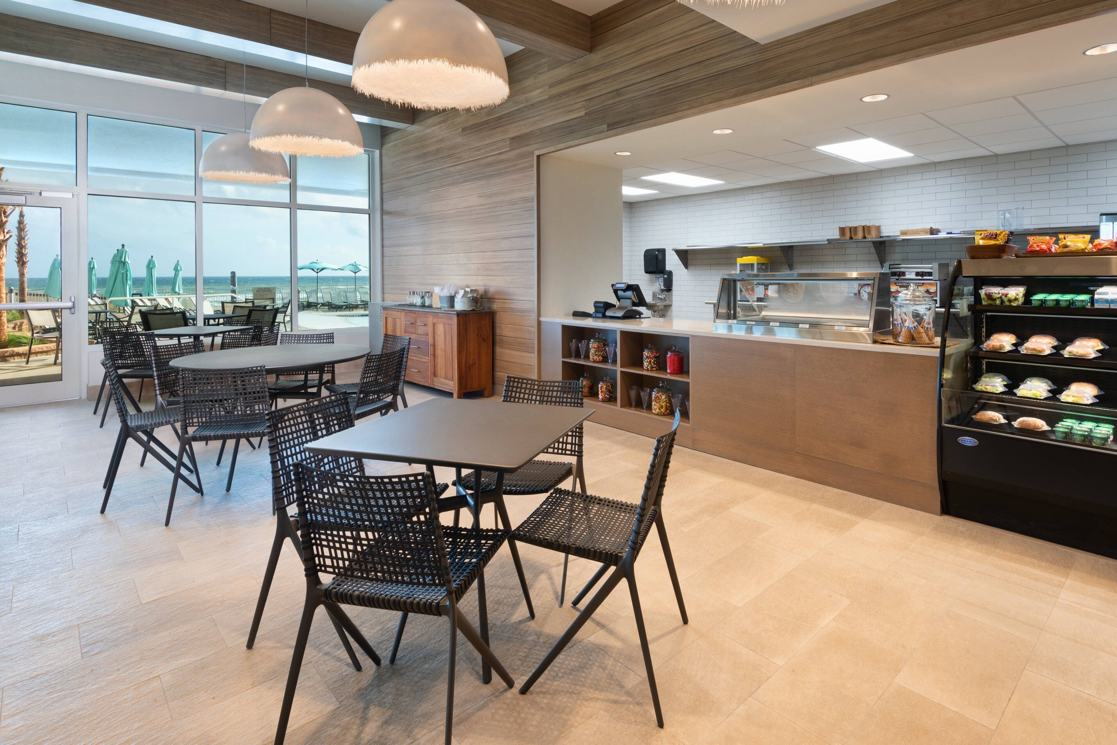 A modern cafe with a view of the beach, featuring a counter with pastries, a seating area with tables and chairs, and a large window overlooking the ocean.