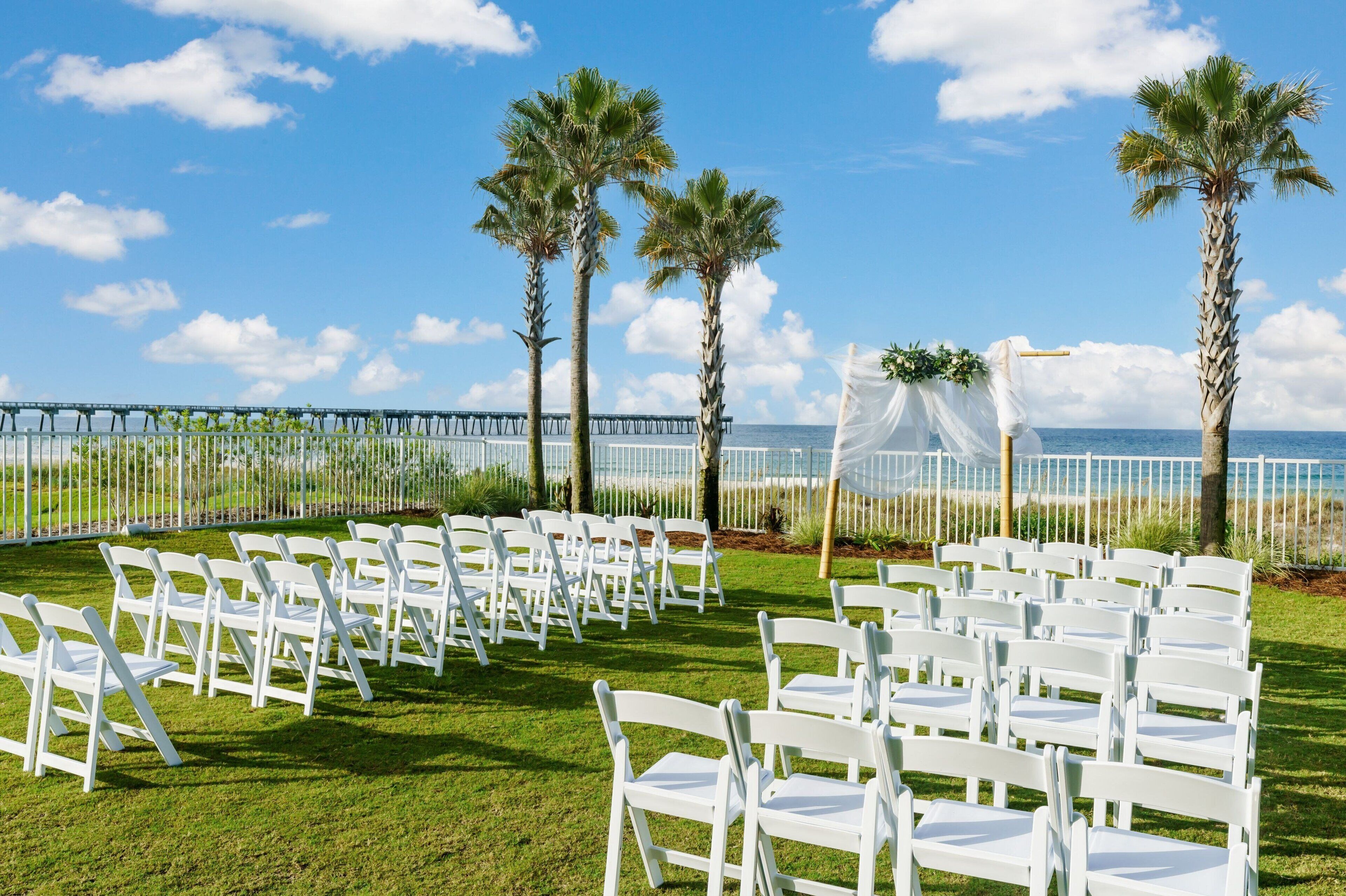 Montaje para una boda al aire libre con sillas blancas dispuestas en filas, palmeras y vista al océano.