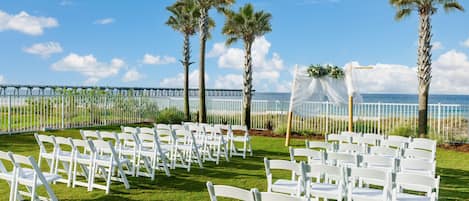 Montaje para una boda al aire libre con sillas blancas dispuestas en filas, palmeras y vista al océano.