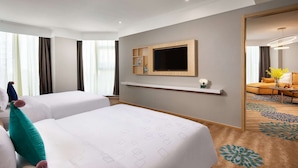Habitación de hotel con dos camas, un televisor montado en la pared, una pequeña estantería y vista de la habitación contigua.