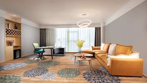 Una habitación de hotel moderna con sofá, escritorio y silla. Cuenta con un ventanal con cortinas, una alfombra de estampado floral y una lámpara colgante.