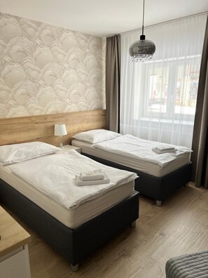 Habitación de hotel con dos camas, cabecero de madera, mesita de noche con lámpara y ventana con cortinas.