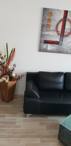 A living room with a black leather sofa, a wooden plant stand with dried plants, and a modern abstract painting on the wall.