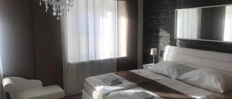 A modern bedroom with a large bed, a bedside table, a chair, a chandelier, and a large mirror.
