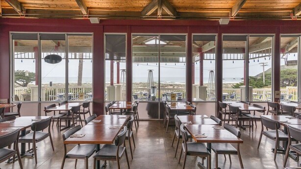 En restaurang med stora fönster som ger utsikt över havet, träbord och stolar samt ett högt tak med synliga takbjälkar.