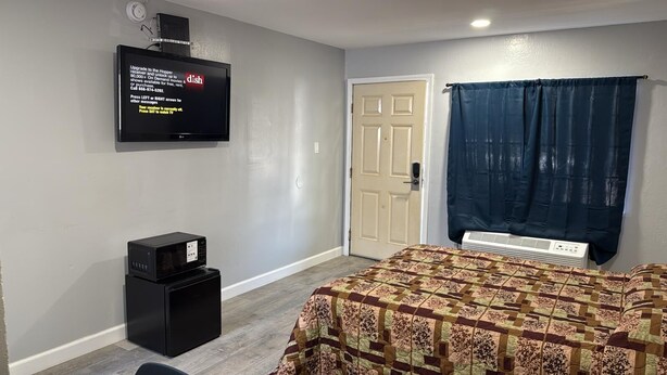 A hotel room with a bed, a television mounted on the wall, a microwave, a door, and a window with curtains. - Palms Motel (Emeryville)