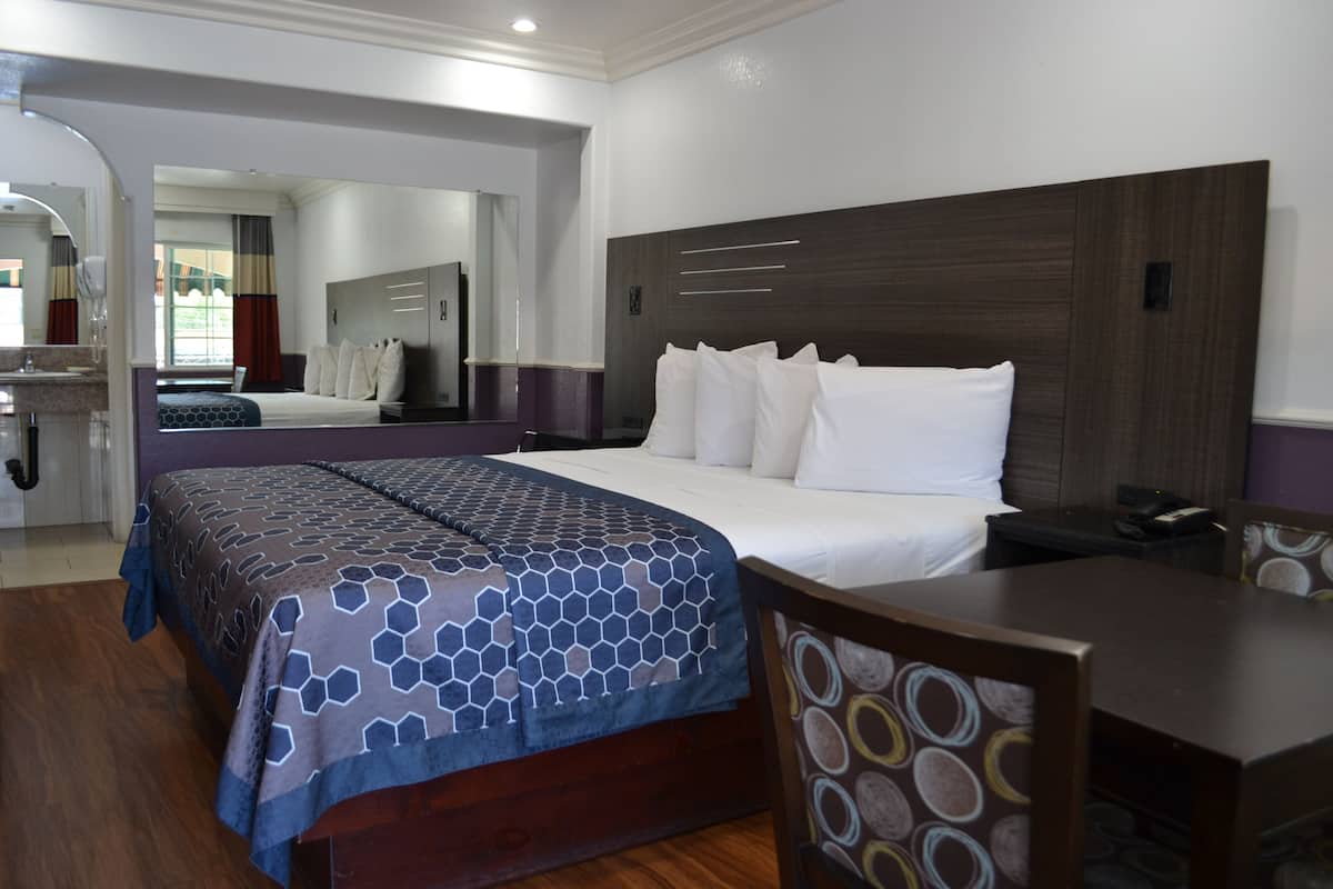 Ein Hotelzimmer mit einem großen Bett, einem Schreibtisch und einem Sessel. An der Wand befindet sich ein Spiegel und ein Fenster mit Vorhängen.