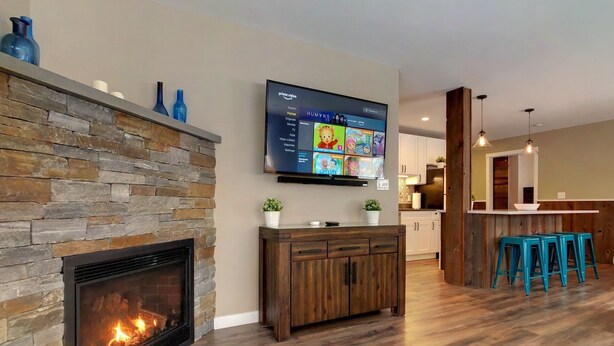 Una sala de estar moderna con chimenea de piedra, un televisor de pantalla plana montado sobre ella, piso de madera y una zona de cocina al fondo.