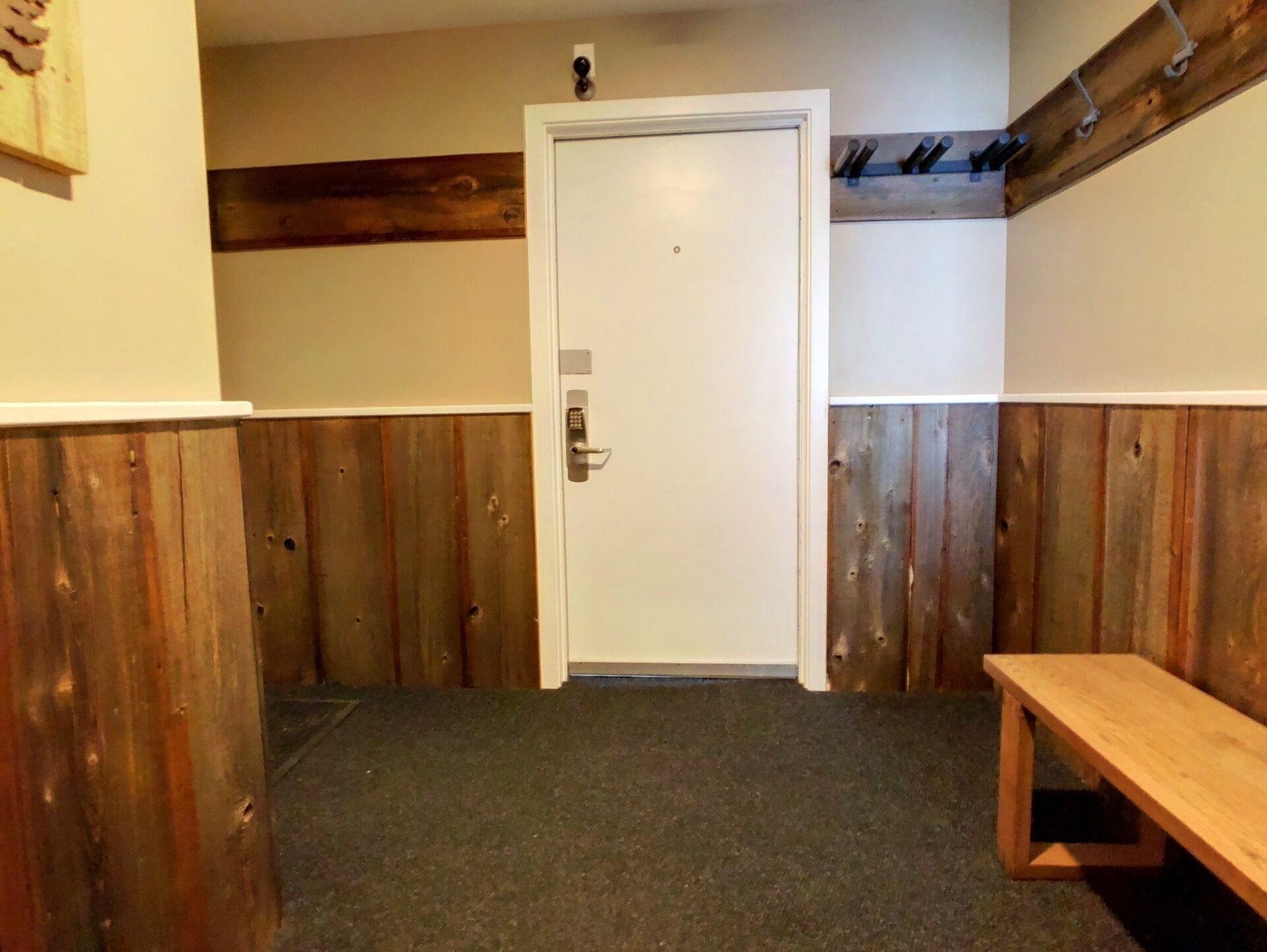 A small, enclosed room with a wooden bench, a door with a keypad lock, and a wooden beam on the ceiling.