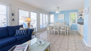 A living room with a blue sofa, a dining table with white chairs, and a kitchen area with blue walls and white cabinetry. - Enjoy Family Memories - Great Outdoor Decks And Backyard; A Few Houses To Beach (Surf City)