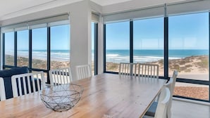 Un espace repas moderne avec une table et des chaises en bois, de grandes fenêtres offrant une vue sur la plage et l’océan.