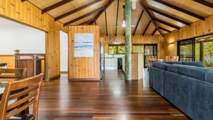 Amplia habitación con paredes de madera y un techo alto, que incluye una zona de comedor con sillas y mesa, un sofá azul y vistas al exterior a través de amplias ventanas.