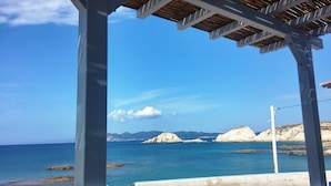 A coastal view with a wooden pergola structure, clear blue sky, and rocky formations in the distance.