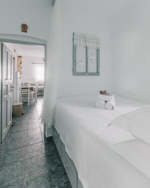 Een slaapkamer met een bed, een klein tafeltje met handdoeken, een raam met gordijn en een gang naar een andere kamer.