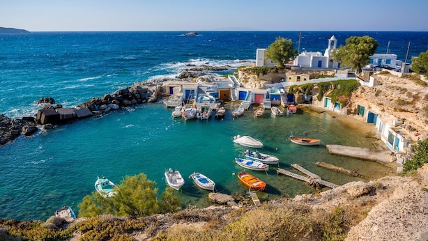A coastal village with traditional white buildings, a rocky shoreline, and boats docked in the clear blue waters.
