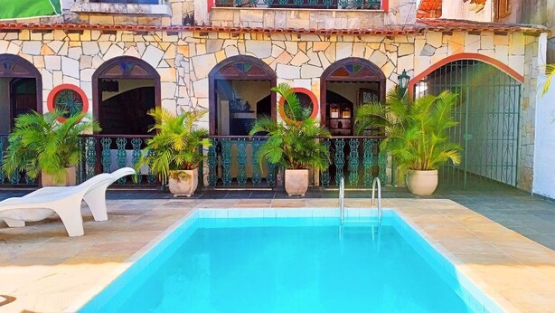 Una piscina con un muro de piedra y ventanas arqueadas, rodeada de plantas en macetas y sillas de descanso.