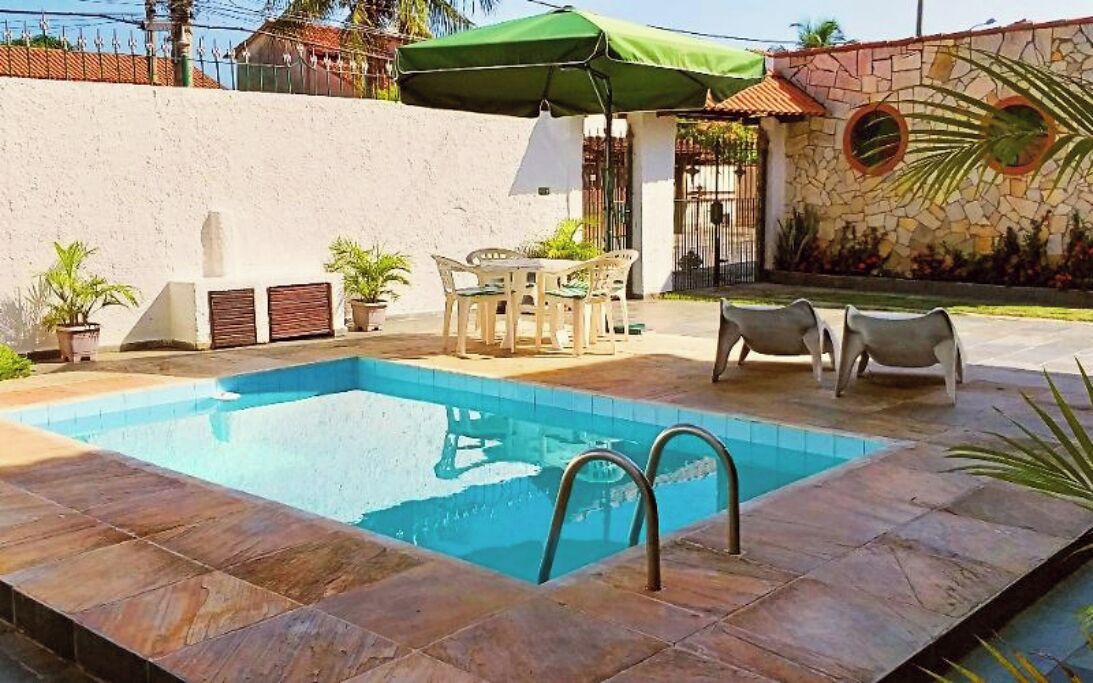 A swimming pool with a stone wall, a green umbrella, and outdoor furniture.