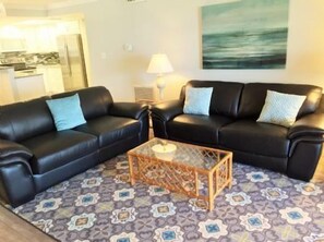 A living room with two black leather sofas, a glass coffee table, patterned rug, and a painting on the wall. - Buttonwood 447- 3 bedroom 2 bath condo with fantastic view of gulf (Siesta Key)