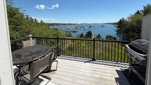 A deck with a grill and a table, overlooking a marina with boats and a clear blue sky. - Enjoy the stunning ocean view from the hot tub at Port! (Southwest Harbor)