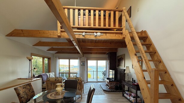 A spacious living area with a high ceiling, wooden beams, a staircase, and a view of the sea through large windows. - Enjoy the stunning ocean view from the hot tub at Port! (Southwest Harbor)