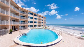 Ein Pool mit Blick auf den Strand, ein mehrstöckiges Gebäude mit Balkonen und ein klarer, blauer Himmel.