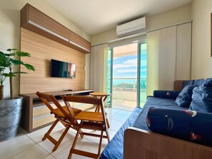 A modern living room with a sofa, a dining table with chairs, a TV mounted on the wall, and a balcony with a view.