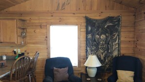 A cozy cabin interior with wooden walls, a dining area with wooden chairs, a table, and a large tapestry.