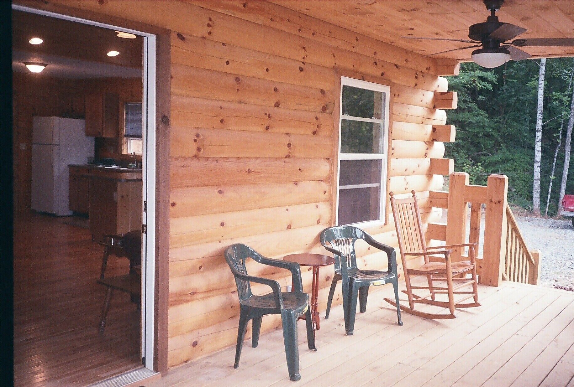 A wooden cabin with a kitchen area visible through an open door, a small table with chairs, and a wooden deck with a rocking chair.