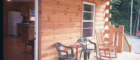 A wooden cabin with a kitchen area visible through an open door, a small table with chairs, and a wooden deck with a rocking chair.