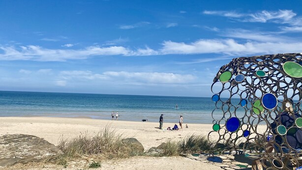 Ein Strand mit blauem Himmel, ruhigem Meer und einer dekorativen Metallskulptur.