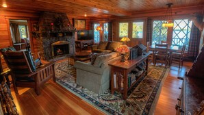 A cozy cabin interior with a stone fireplace, wooden furniture, and a large window providing a view of the outdoors. - HISTORIC LAKE PLACID WATERFRONT GREAT CAMP-6 BR- Each w/private full bath. (Lake Placid)
