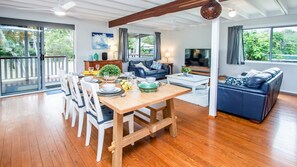 Uma sala de estar ampla com uma mesa de jantar de madeira, cadeiras brancas, um sofá azul e uma televisão.