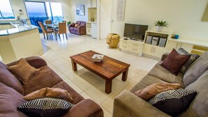 Un vaste espace de vie comprenant une table basse en bois, deux canapés avec des coussins à motifs, un téléviseur et un coin cuisine équipé d’un réfrigérateur et de placards.