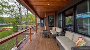 Una terraza de madera con mobiliario exterior, ventanas amplias y vista a un lago.
