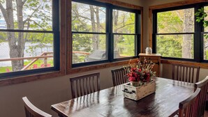 Un comedor con mesa y sillas de madera, ventanas amplias con vistas a árboles y una terraza, y un florero con flores sobre la mesa.